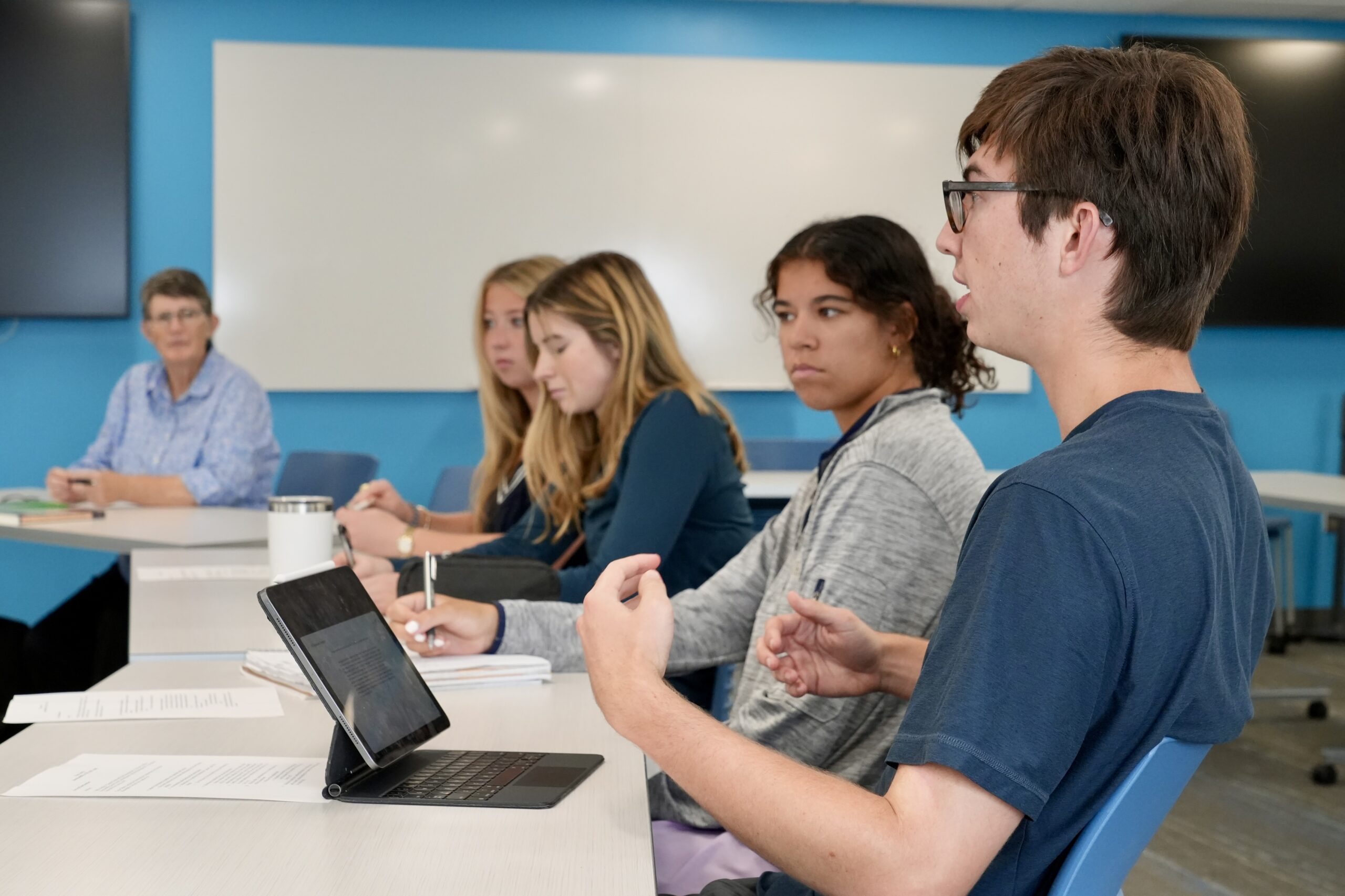 Male student on right gestures and speaks while Professor Margie Pfeil and three female students listen in classroom with light blue walls.